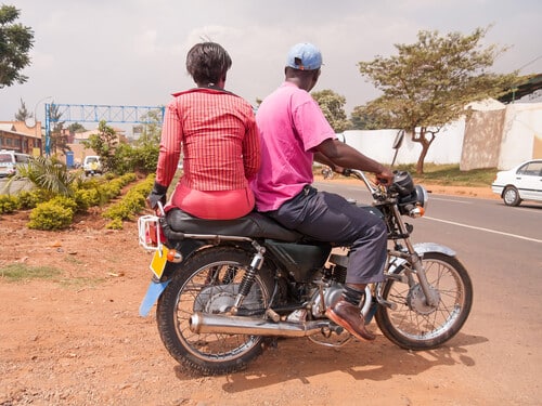 Woman and man sit back on motor bike on roadside. Kampala, Uganda.